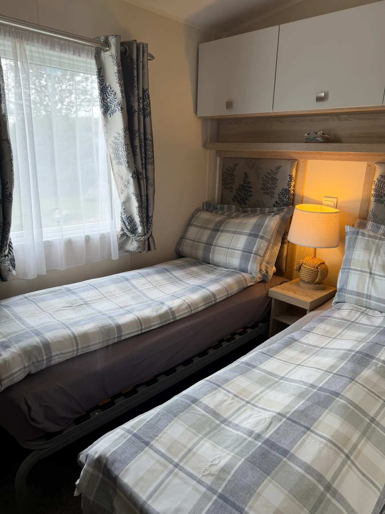 Twin bedroom inside Tern 32 with two single beds in blue-and-white plaid bedding, a rope bedside lamp, overhead cupboards and patterned curtains