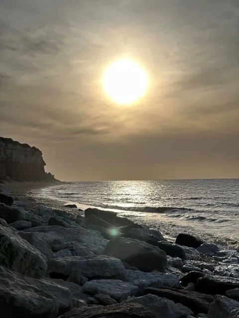 The sun setting over the sea through hazy cloud, with cliffs in silhouette and rocks on the shore