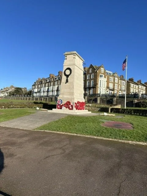 A memorial green with a pale stone memorial, a wreath, an American flag and old buildings behind