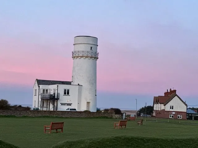 A white round old tower with a railed gallery beside a green with benches under a pink and lilac sky