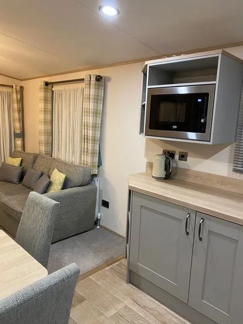 The kitchen inside Windsor 30 with grey units, a wooden worktop, a built-in microwave and a kettle, with the lounge sofa beyond